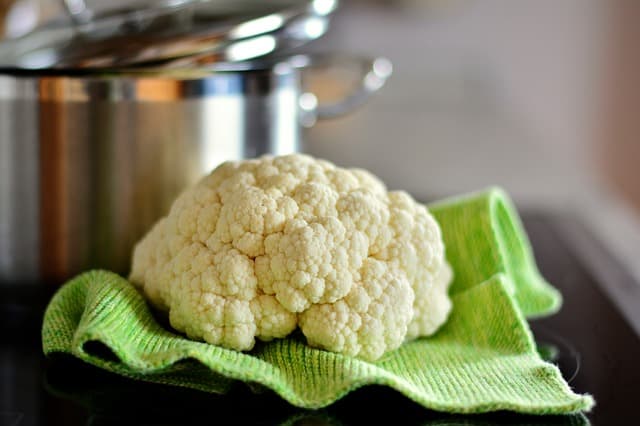 A head of raw cauliflower on a green hand towel with a silver stock pot in the background.